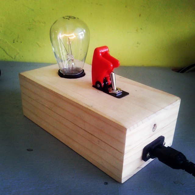 Archivo jpg de una fotografía de un foco incandescente donde se puede apreciar su filamento ardiendo, colocado en un soquet en una caja de madera y un switch eléctrico de palanca con cubierta color roja